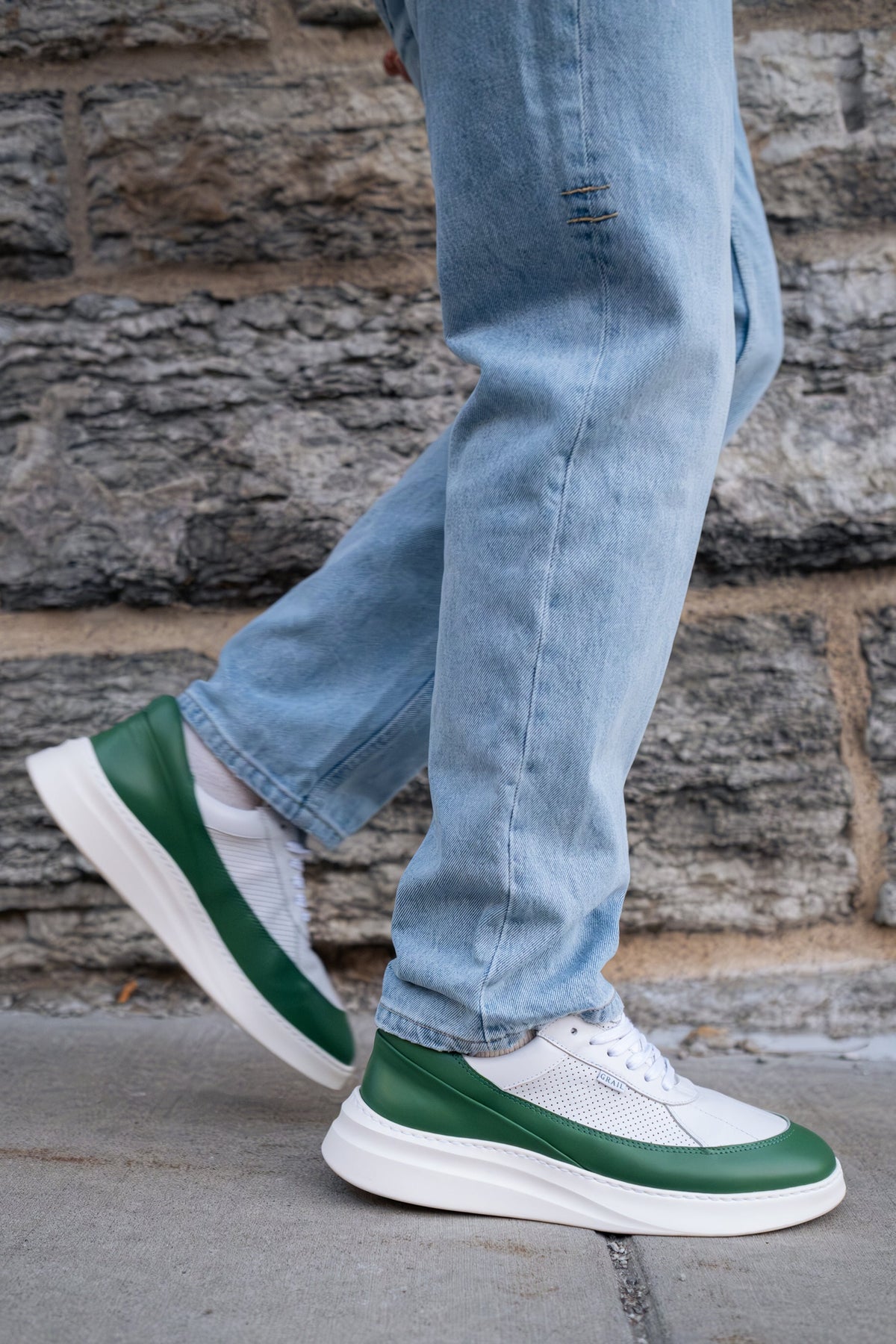 White and green low-top sneakers worn with light-wash blue jeans, shown from the side as a person walks against a gray stone wall.